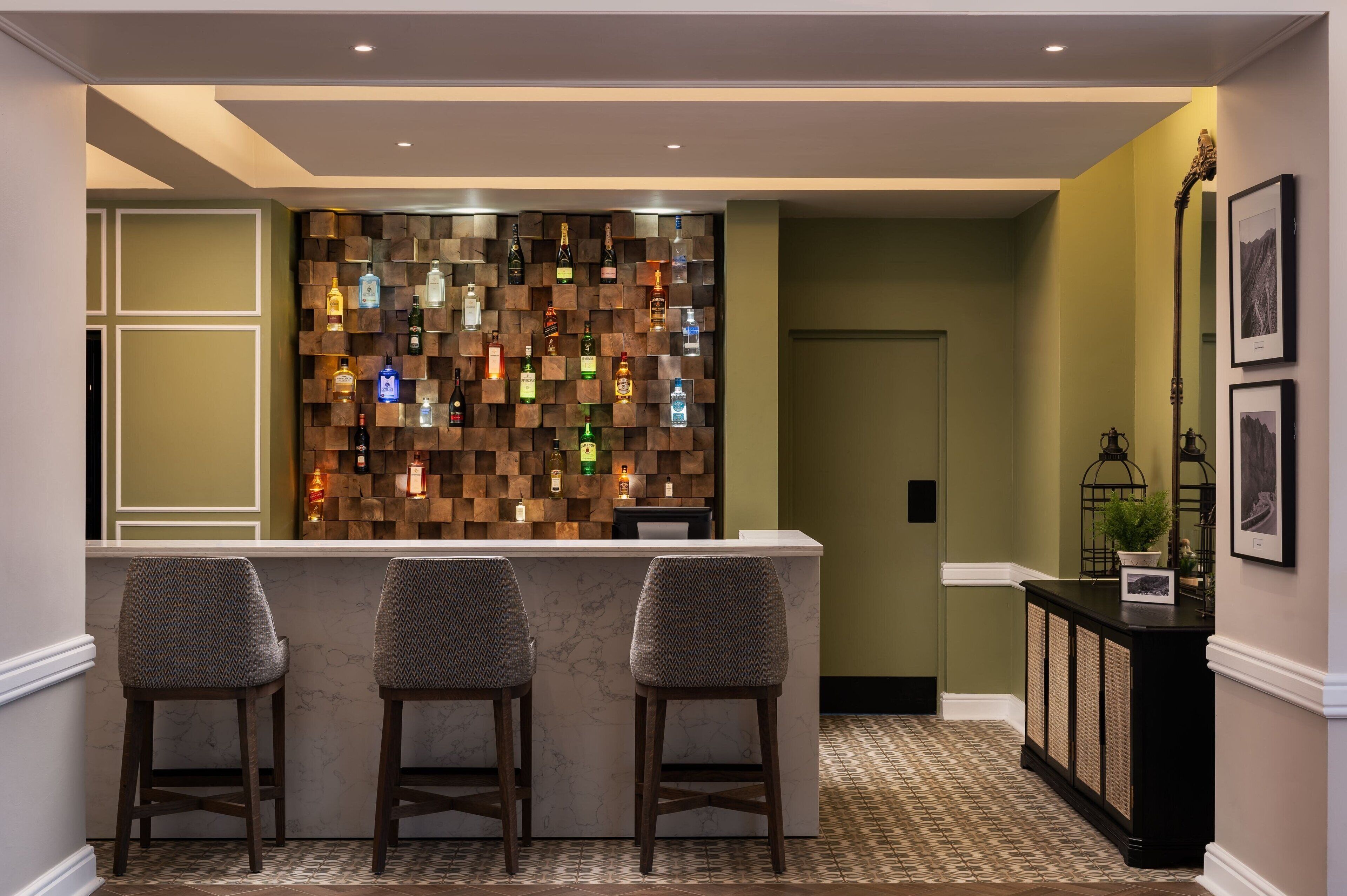 Een bar met een houten wand waarop drankflessen staan, een marmeren toonbank en drie barstoelen.