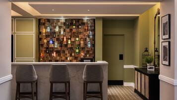 Een bar met een houten wand waarop drankflessen staan, een marmeren toonbank en drie barstoelen.