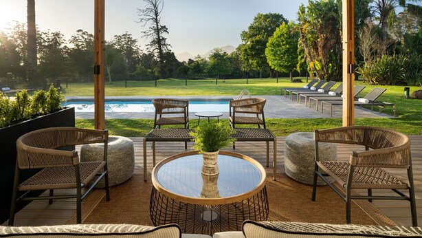 An outdoor patio with wicker furniture, a round table with a vase, and a view of a swimming pool and greenery. - Protea Hotel by Marriott George King George (George)
