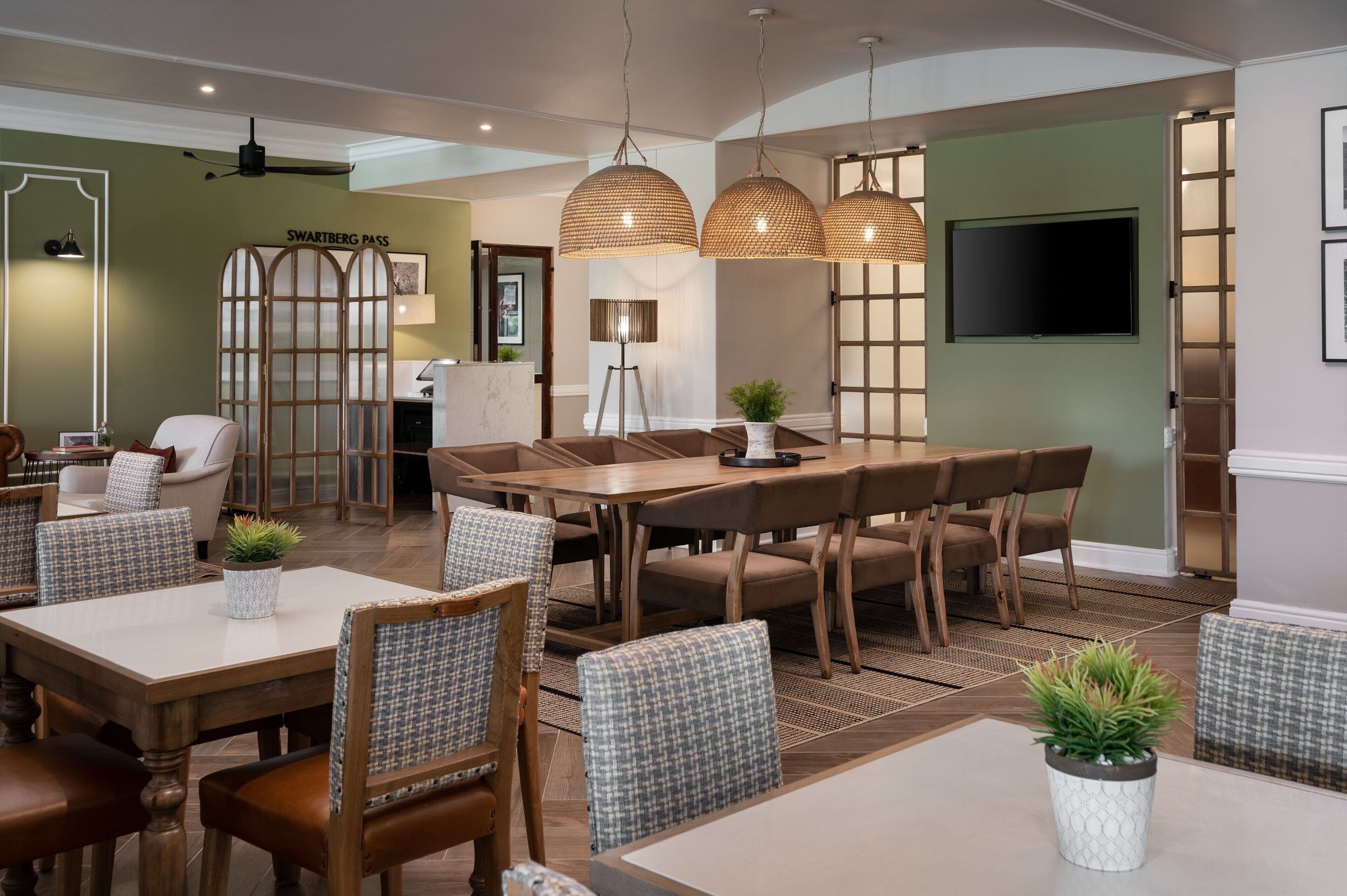 A dining area with a large table and chairs, a television mounted on the wall, and a decorative plant on the table.