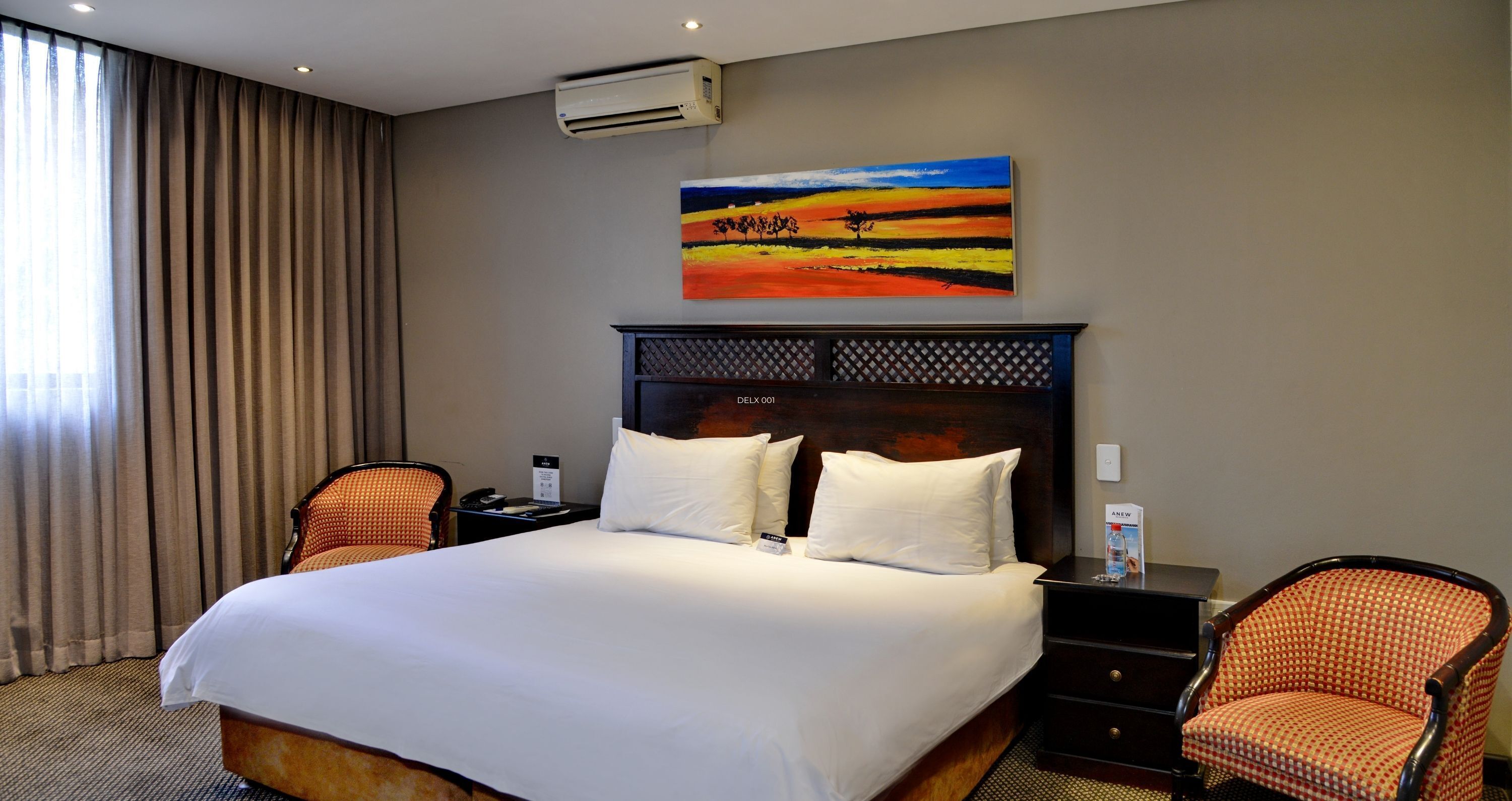 A hotel room with a large bed, two nightstands, a wall-mounted air conditioner, and a painting above the bed.