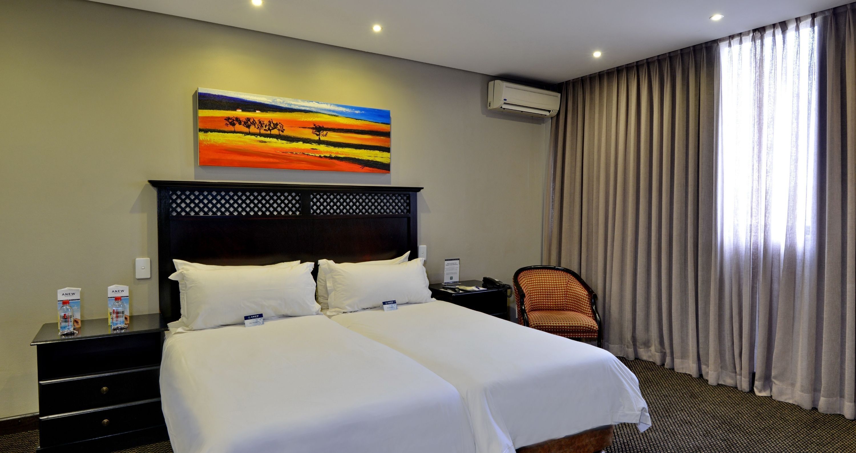 A hotel room with a large bed, two bedside tables, an air conditioning unit, a chair, and a painting on the wall.