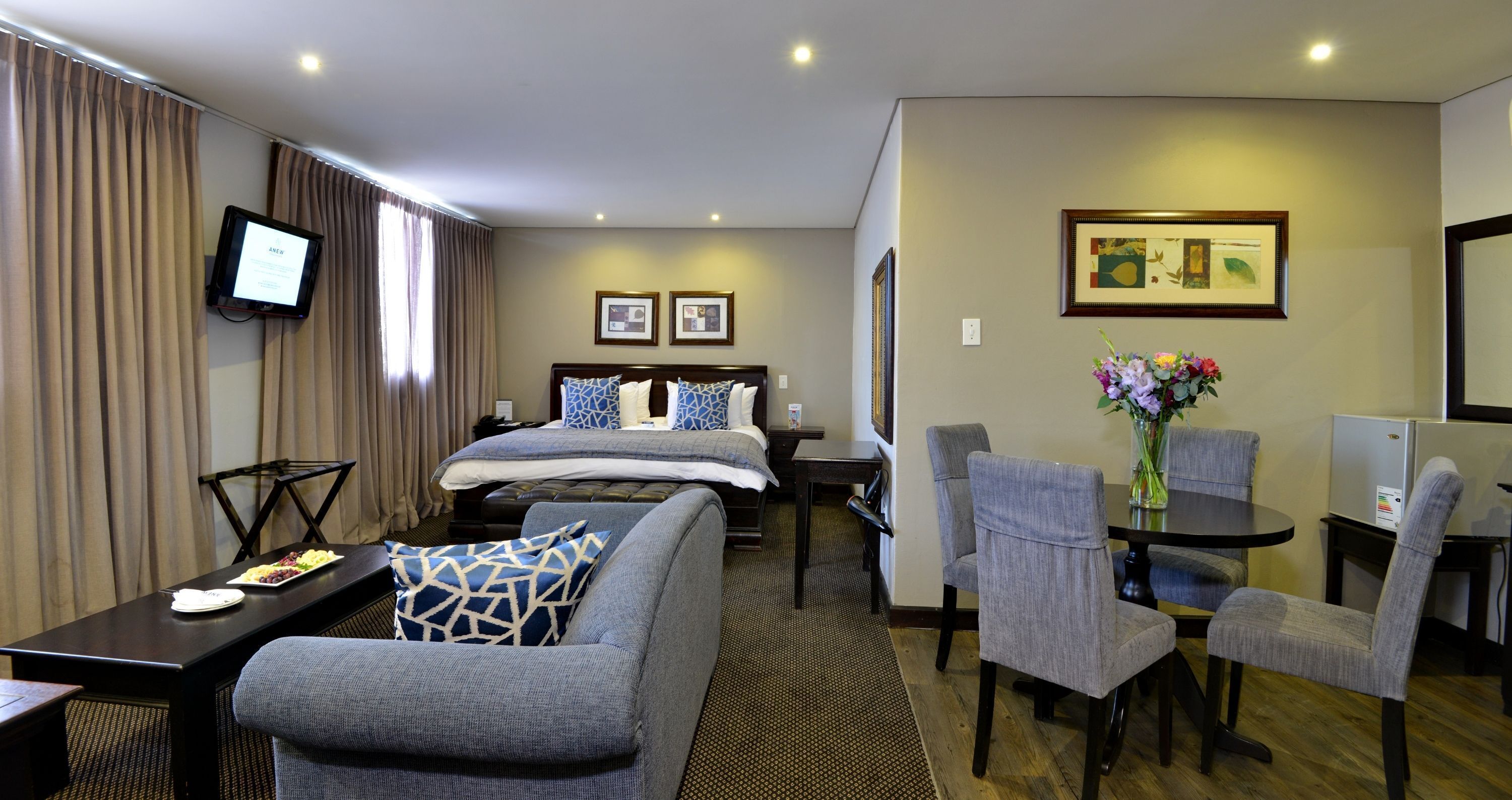 A hotel room with a bed, a desk with a chair, a dining table with chairs, a TV mounted on the wall, and framed pictures on the wall.