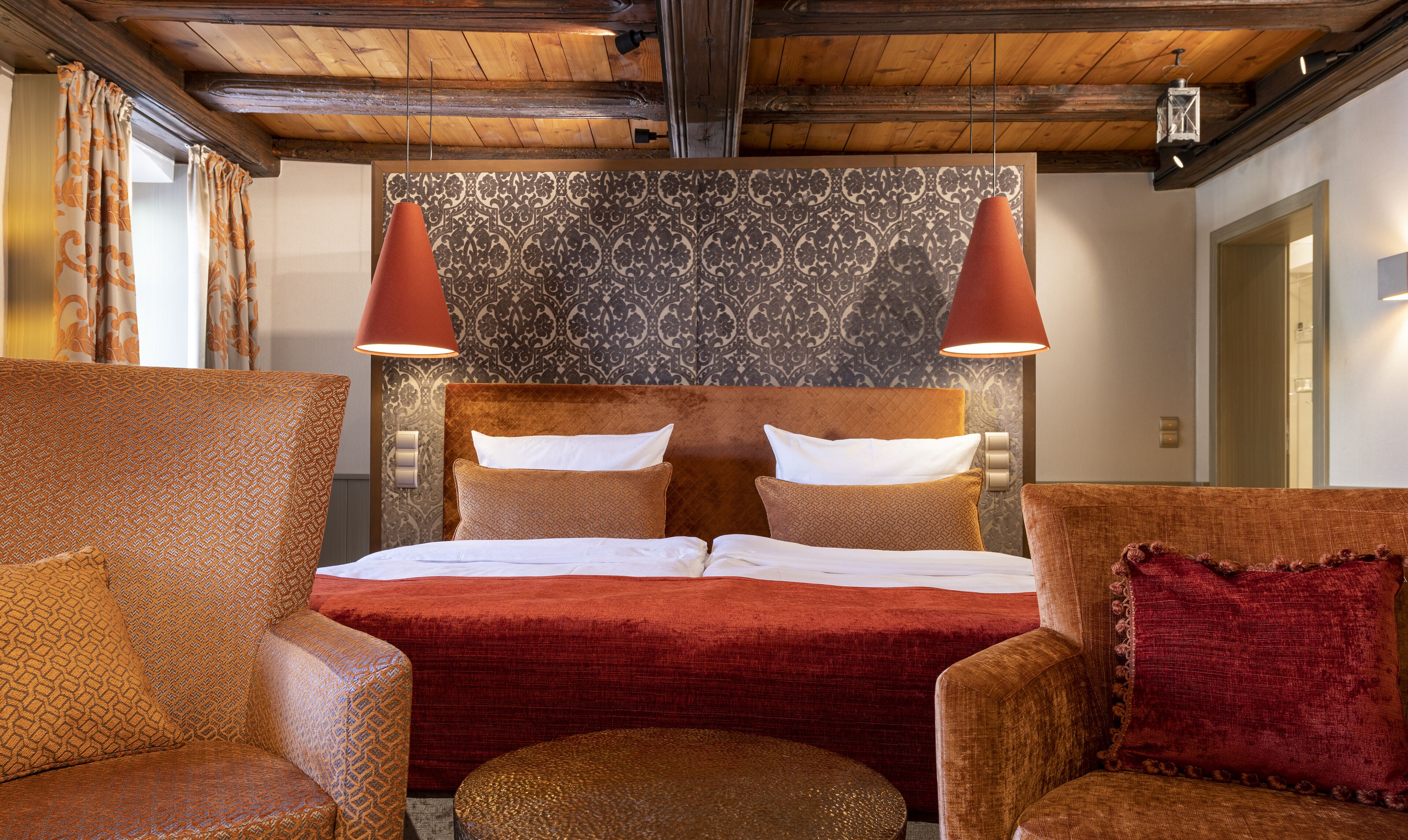 Una habitación de hotel con una cama grande, dos sillones, un puf redondo y un papel tapiz estampado.
