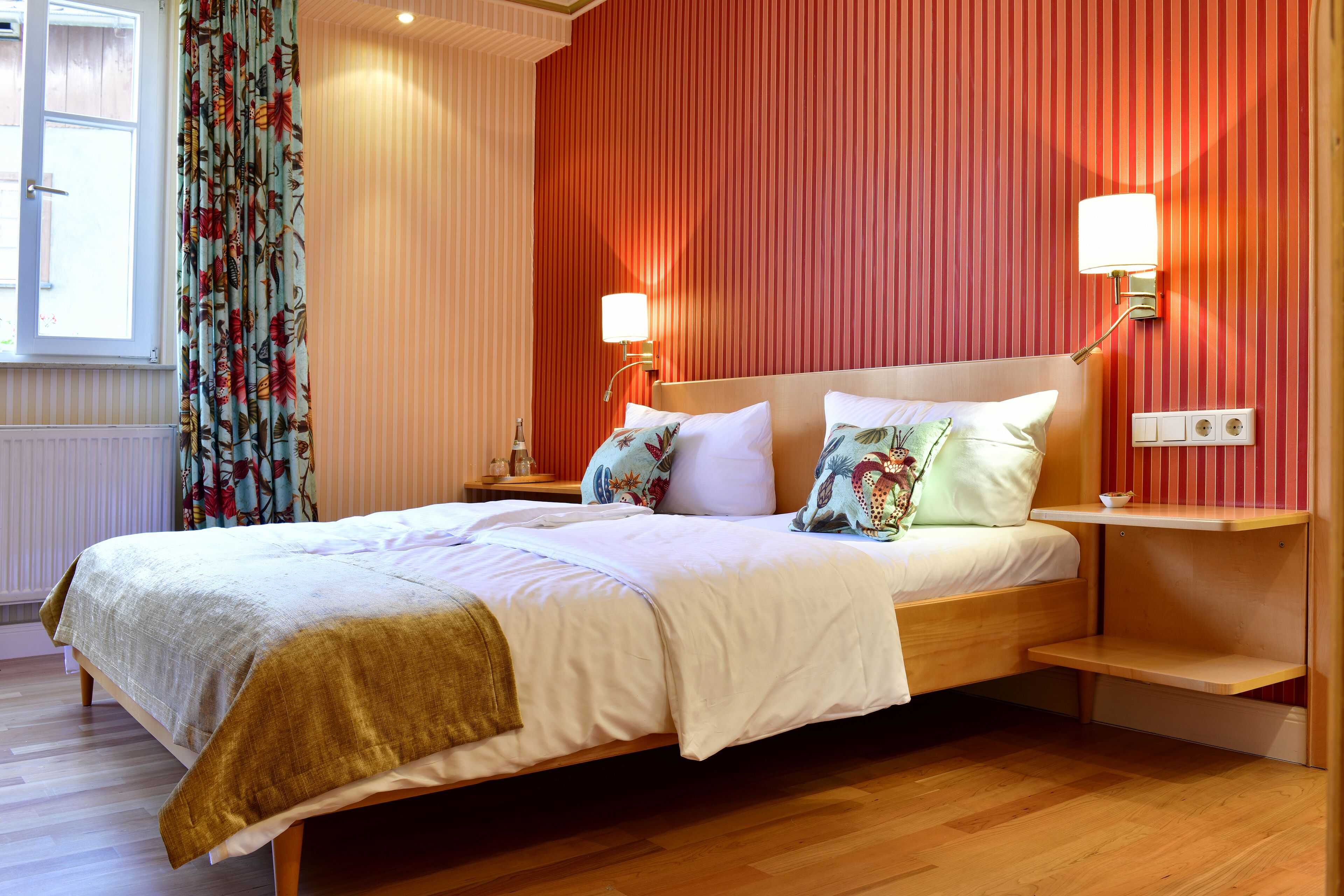 Una cama bien hecha con cubrecama blanco y dorado, dos mesitas de noche con lámparas y una ventana con cortinas.