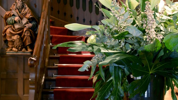 Una escalera con peldaños alfombrados en rojo, barandilla de madera y una estatua a la izquierda. Una gran planta en maceta con hojas verdes está sobre una mesita junto a la escalera.