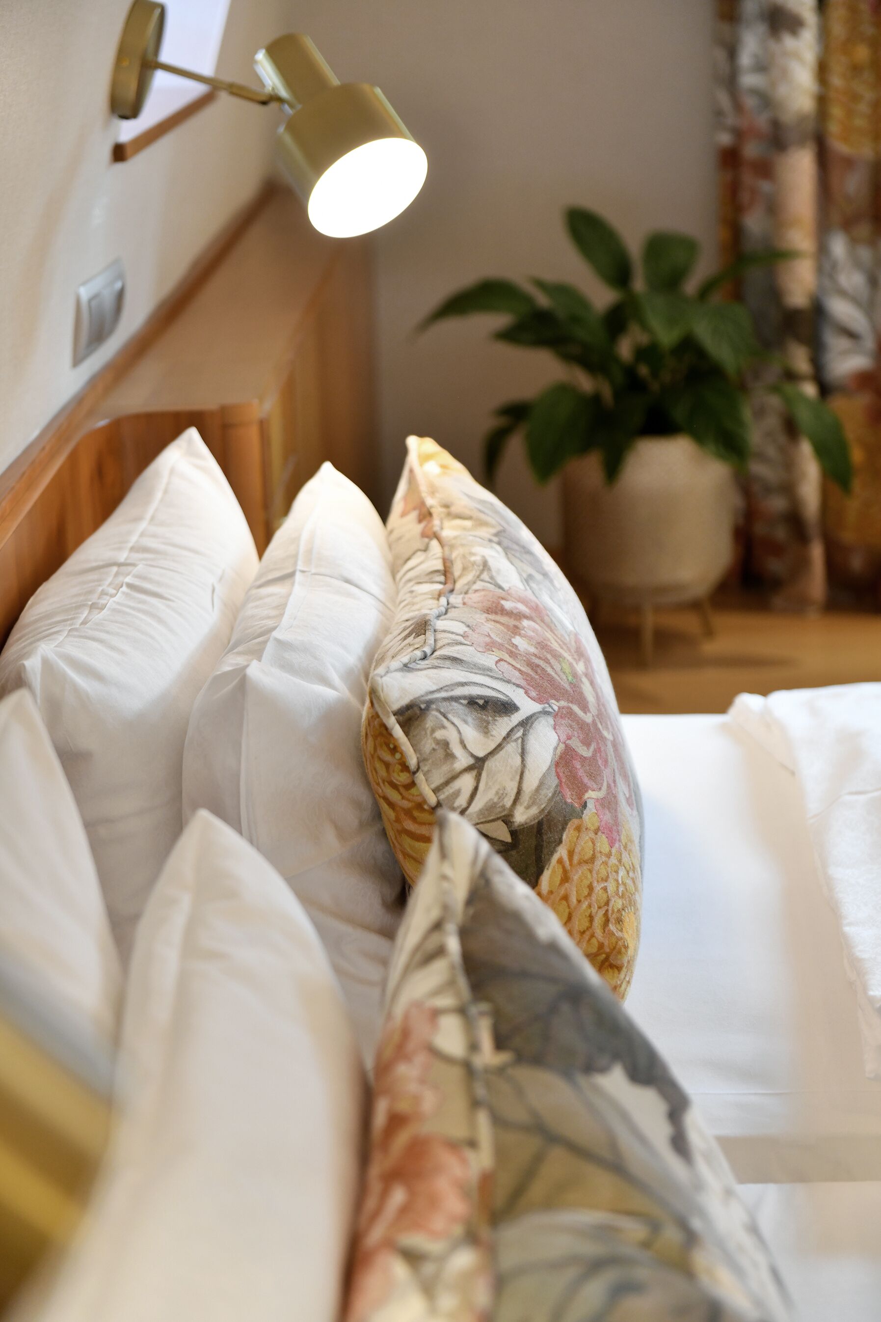 Una cama bien hecha con almohadas blancas y estampadas, una lámpara de pared y una planta en maceta al fondo.