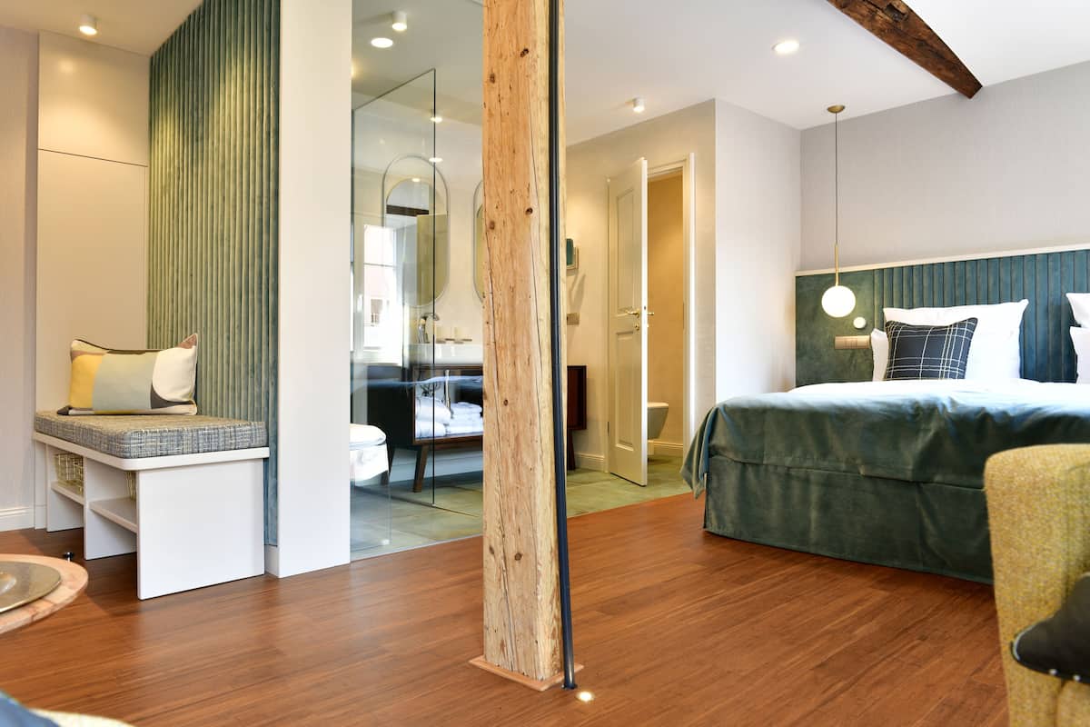 Un dormitorio moderno con suelo de madera, una cama con cabecero verde, una zona de estar con un banco y un espejo grande.