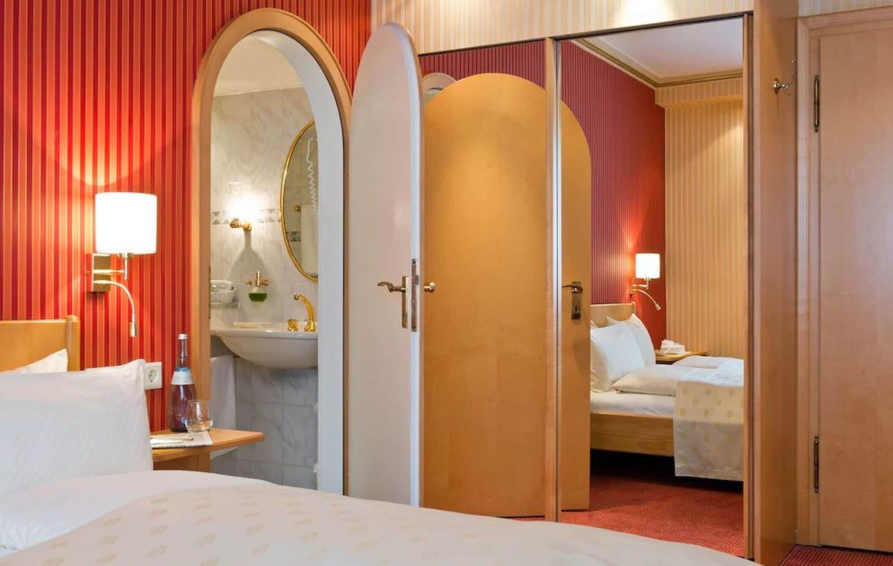 A hotel room with a bed, a bedside table, a lamp, a mirror, a sink, and a door leading to another room.