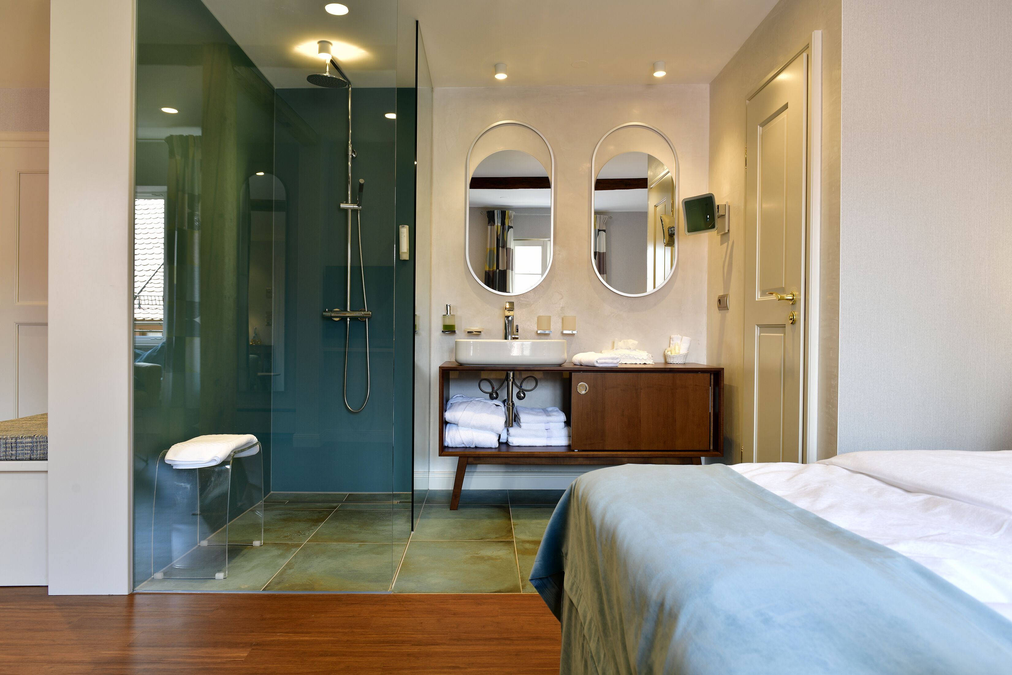 A modern bathroom with a glass shower enclosure, a vanity with a sink and mirror, and a bed with a blue bedspread.
