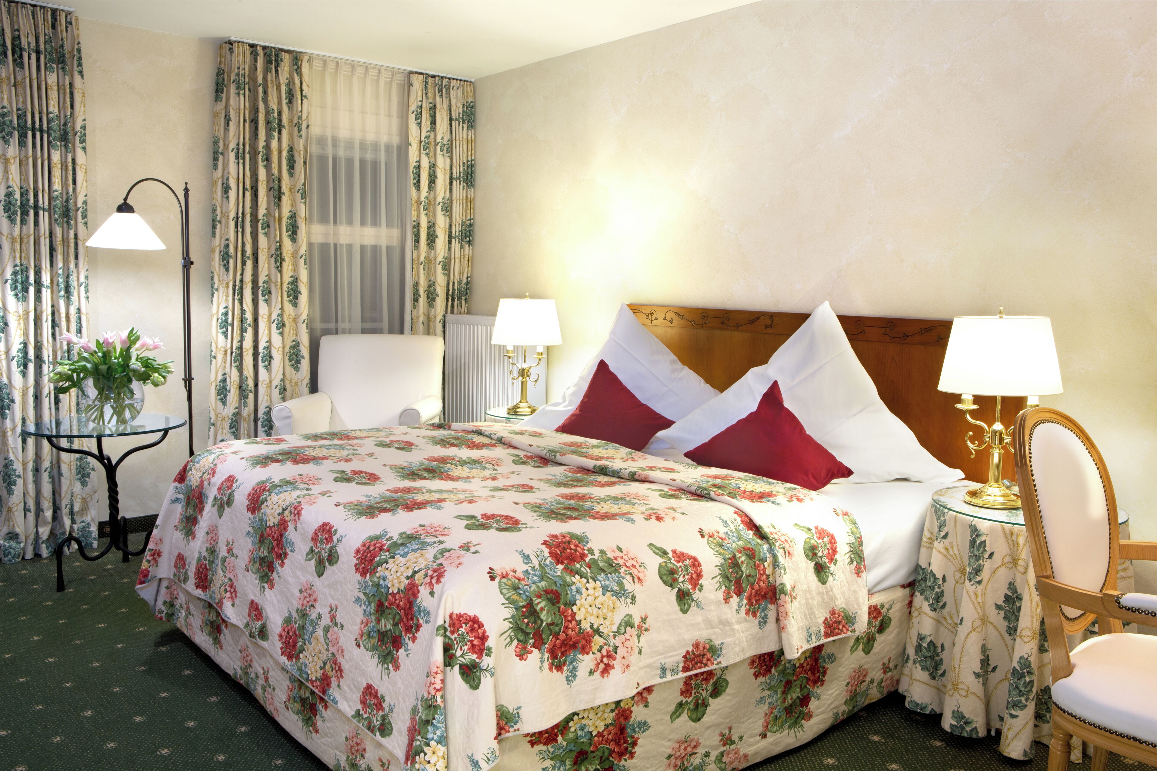 A hotel room with a floral bedspread, two bedside lamps, a chair, and a small table with a plant.