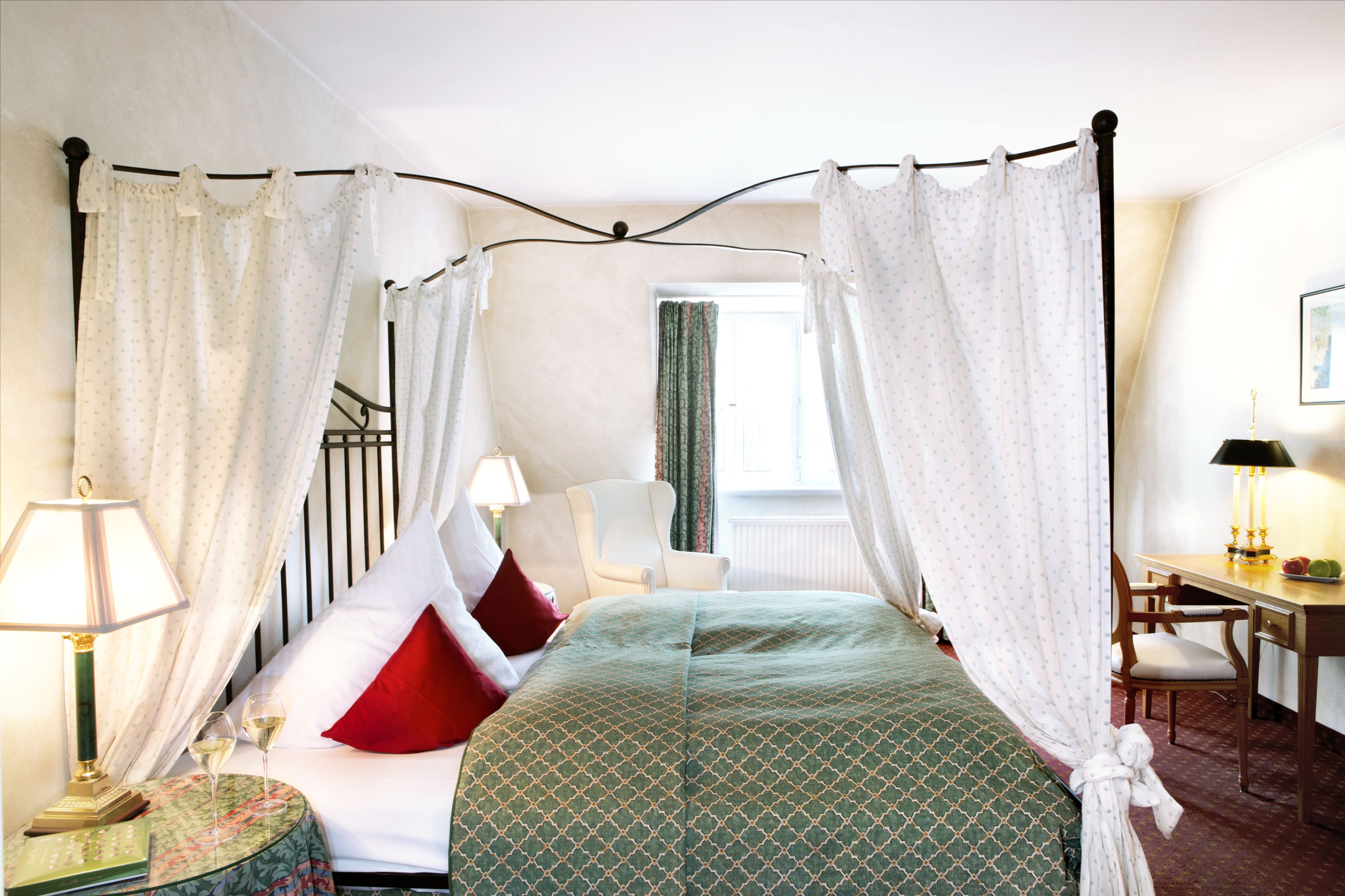 A four-poster canopy bed with a green patterned bedspread, two red pillows, and a bedside table with a lamp.