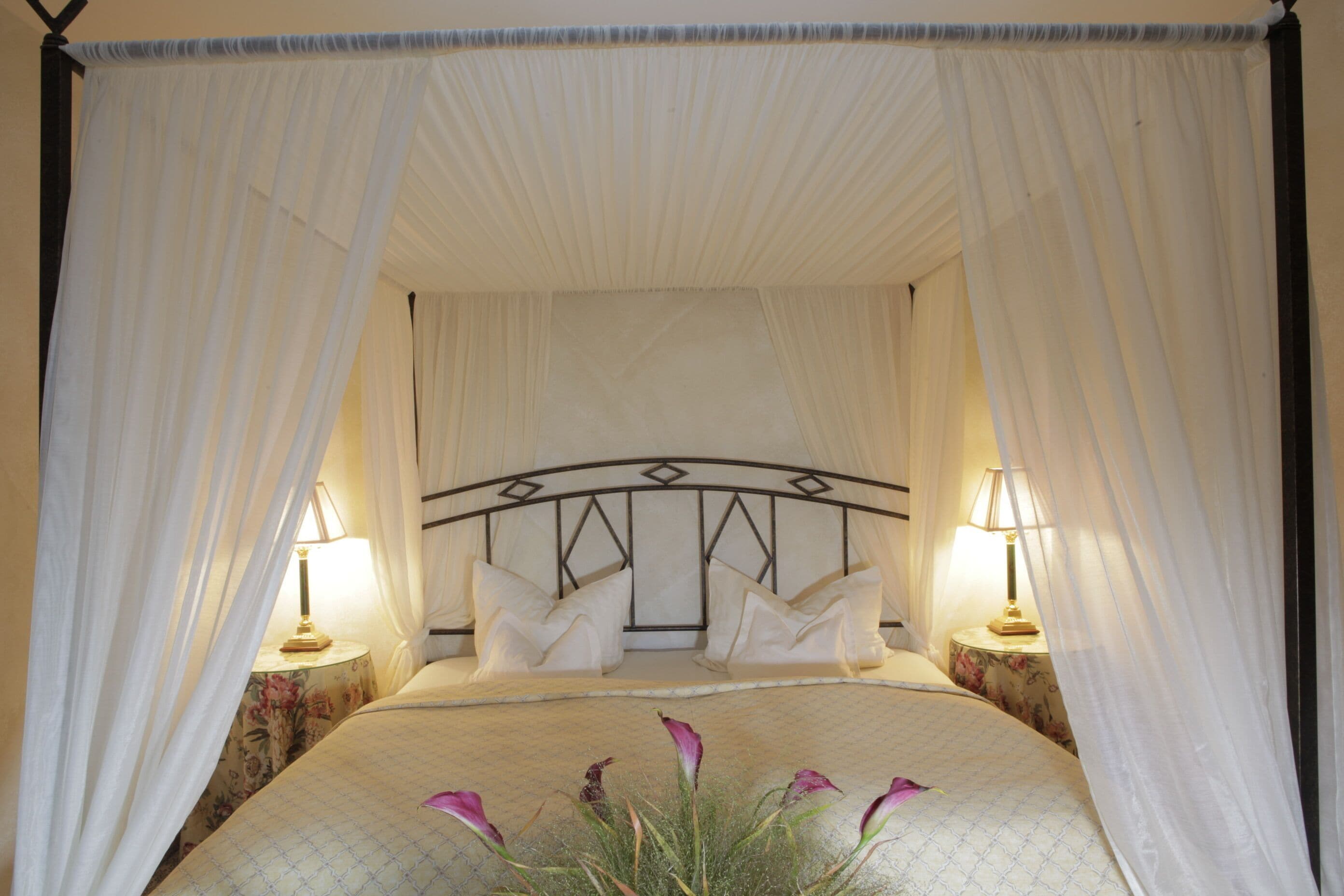 A bed with a canopy, two bedside tables with lamps, and a floral-patterned tablecloth.
