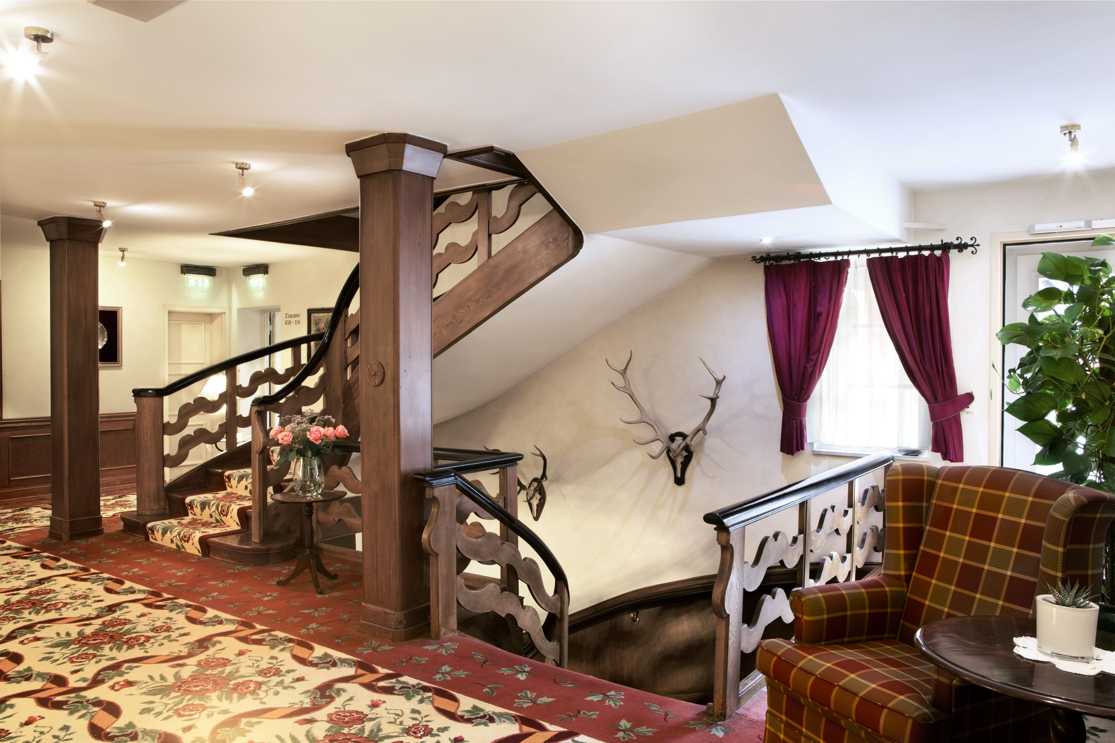 Una imponente escalera con barandales ornamentados, un suelo con motivos florales, un sillón mullido y un cráneo de ciervo en la pared.