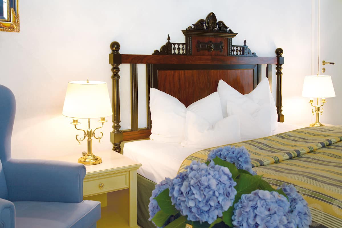 Een hotelkamer met een houten hoofdeinde, witte beddengoed, een blauwe fauteuil, twee nachtkastjes met lampen, en een bos blauwe bloemen.