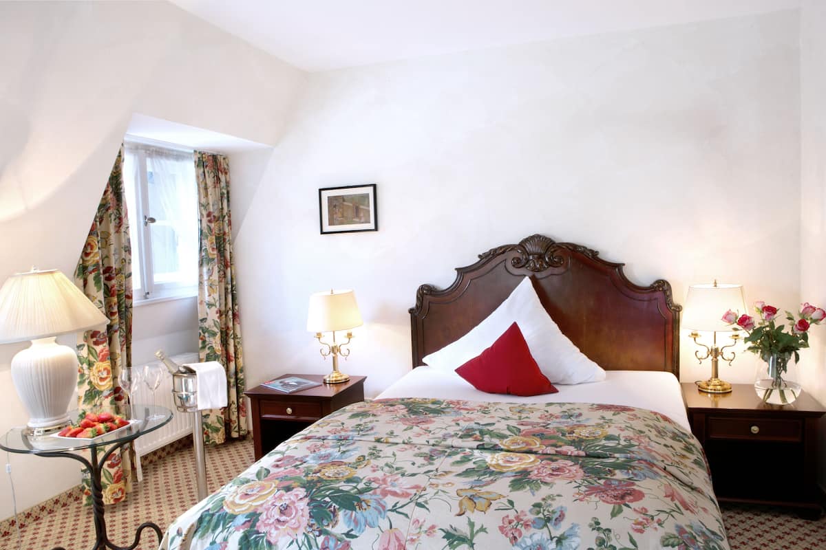 Hotelkamer met groot bed, bloemenprint beddegoed, twee nachtkastjes met lampen, een tafeltje met lamp en fruit, en een raam met gordijnen.