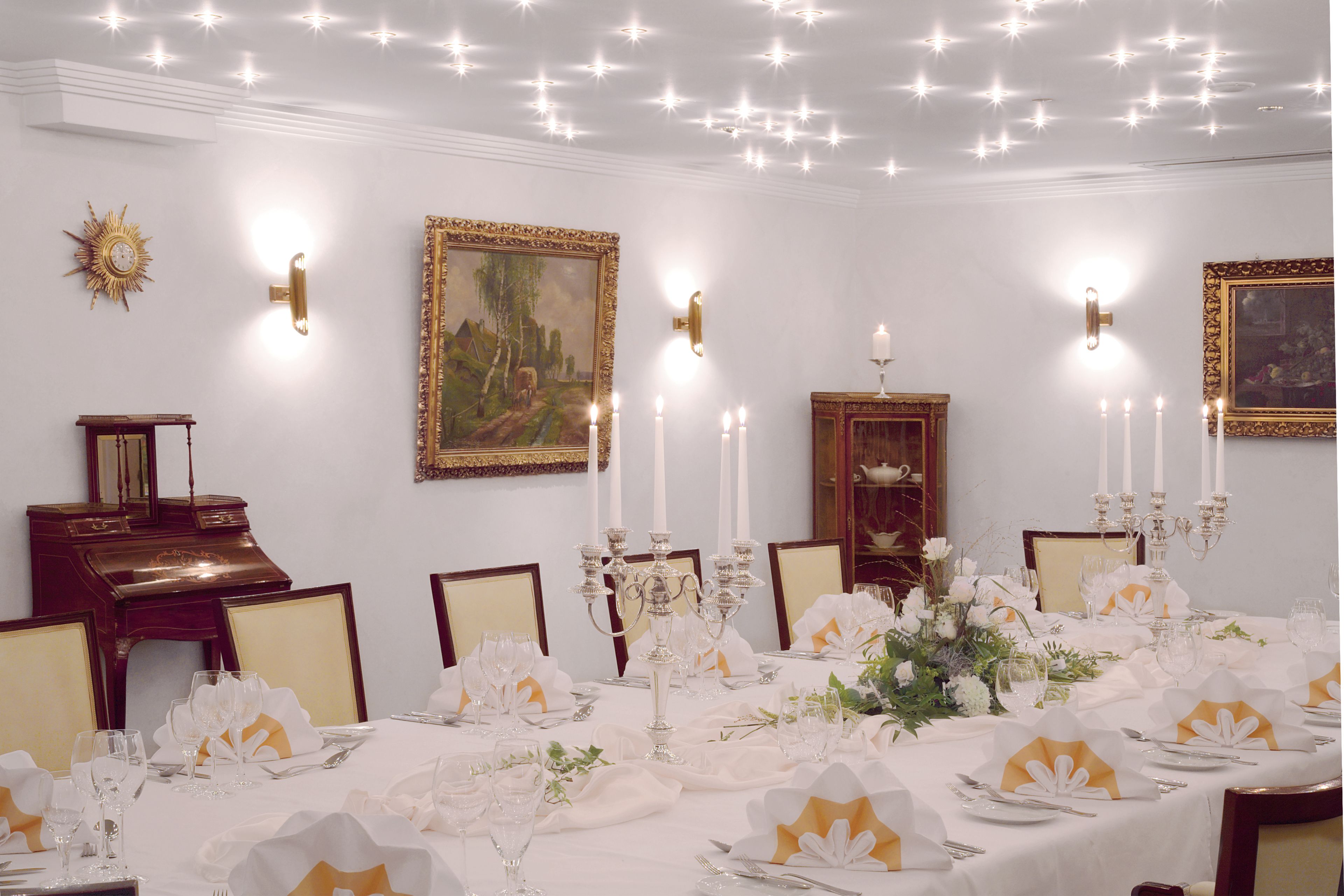 A formal dining room with a long table set for a meal, surrounded by wall-mounted sconces and framed artwork.