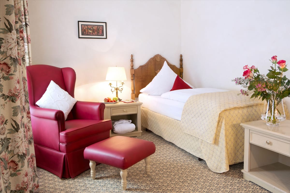 Hotelkamer met een rode fauteuil, een bed met witte beddengoed, een nachtkastje met een lamp en een vaas met bloemen.
