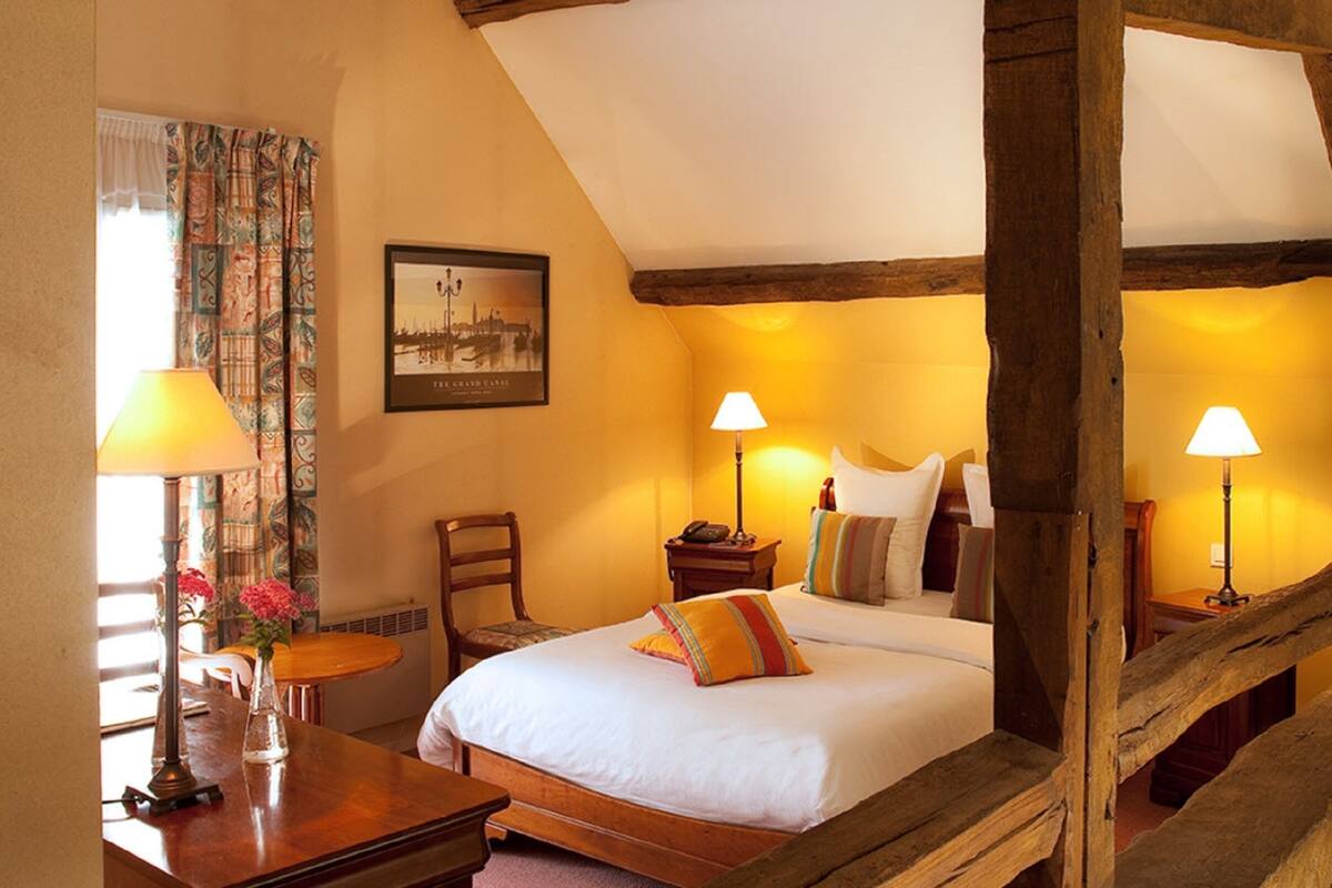 Une chambre à coucher avec un lit à baldaquin, deux lampes de chevet, une chaise, une petite table avec une lampe et un tableau encadré au mur.