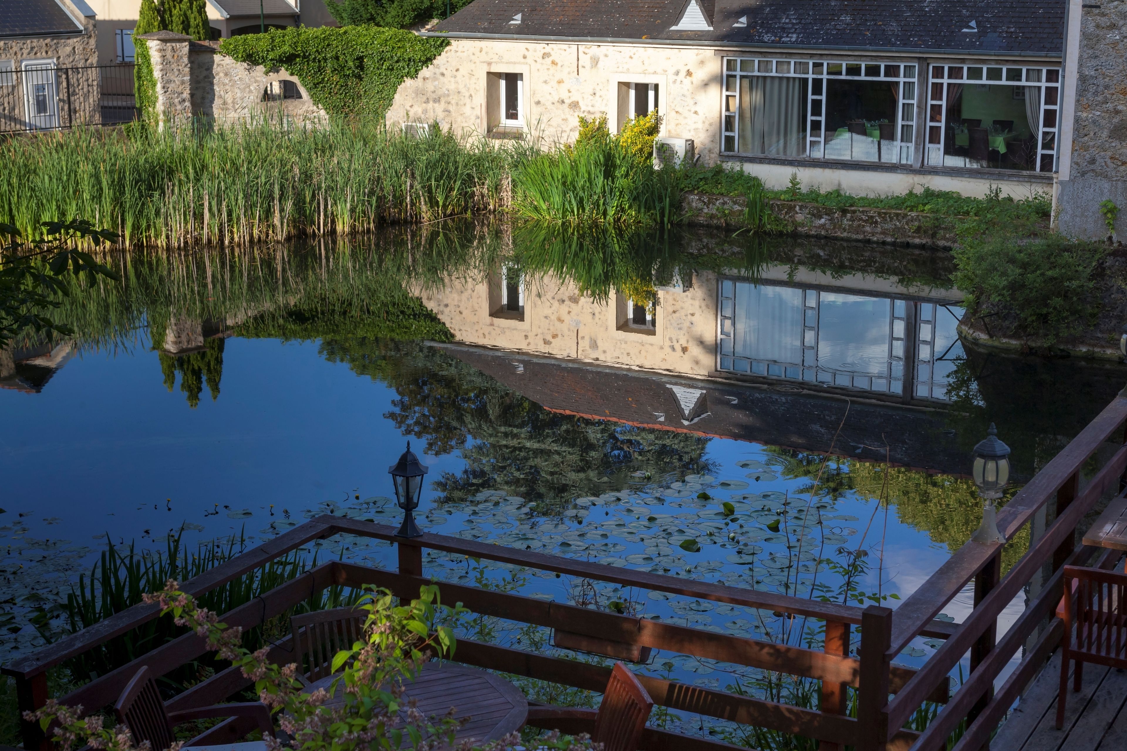 A serene riverside view with a wooden deck, stone buildings, and a clear reflection in the water.
