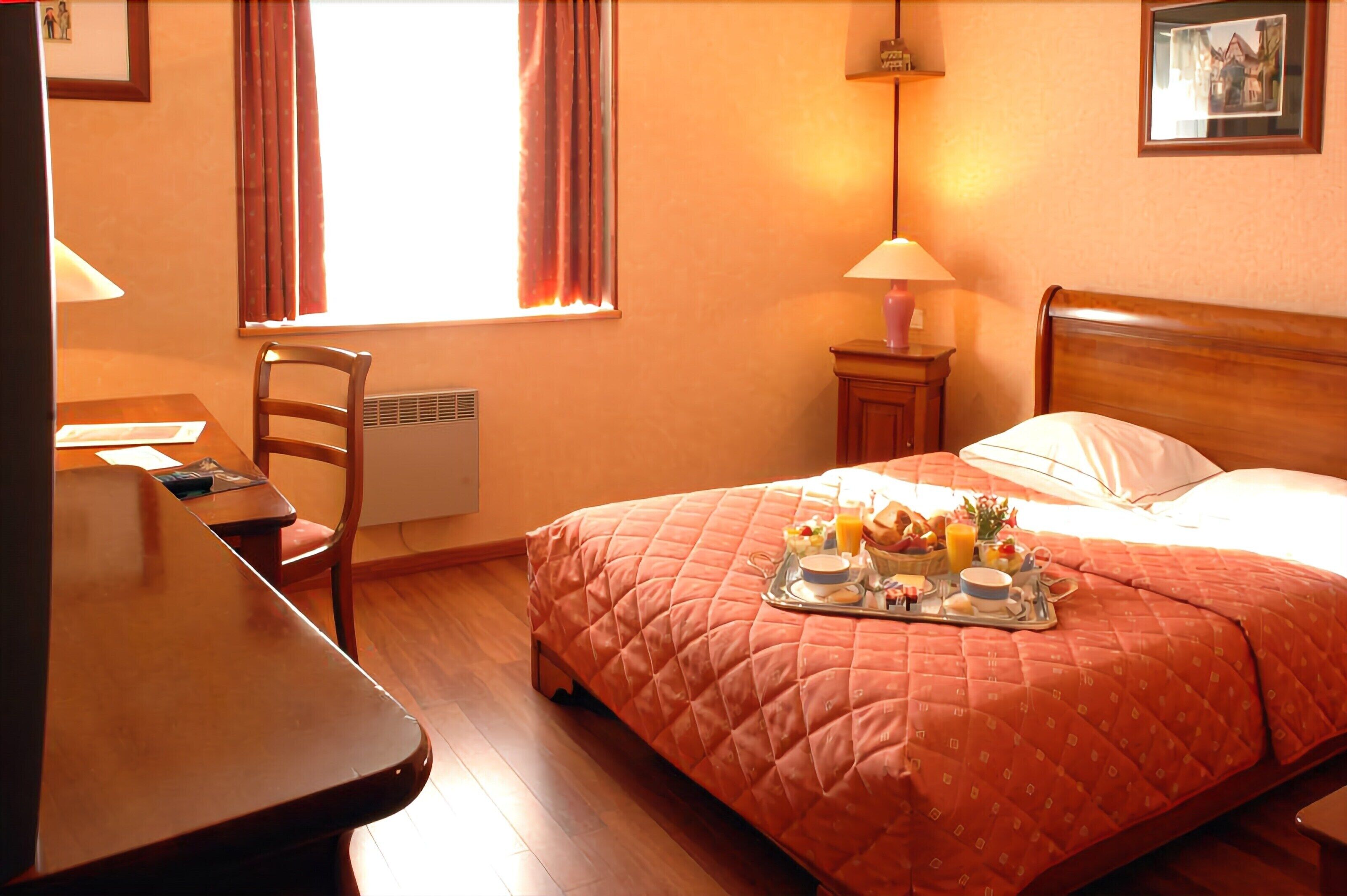 A hotel room with a bed, a desk, a chair, a nightstand with a lamp, and a window with curtains.