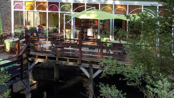 A riverside restaurant with outdoor seating, a wooden deck, and a glass enclosure.
