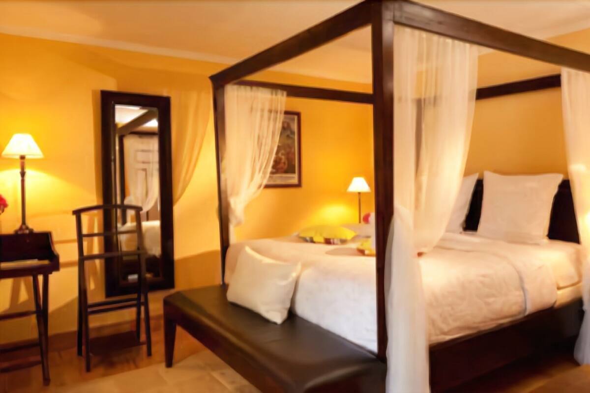 Un lit à baldaquin avec des draps blancs et un dais, un banc en bois, un bureau avec une chaise et deux lampes de chevet.