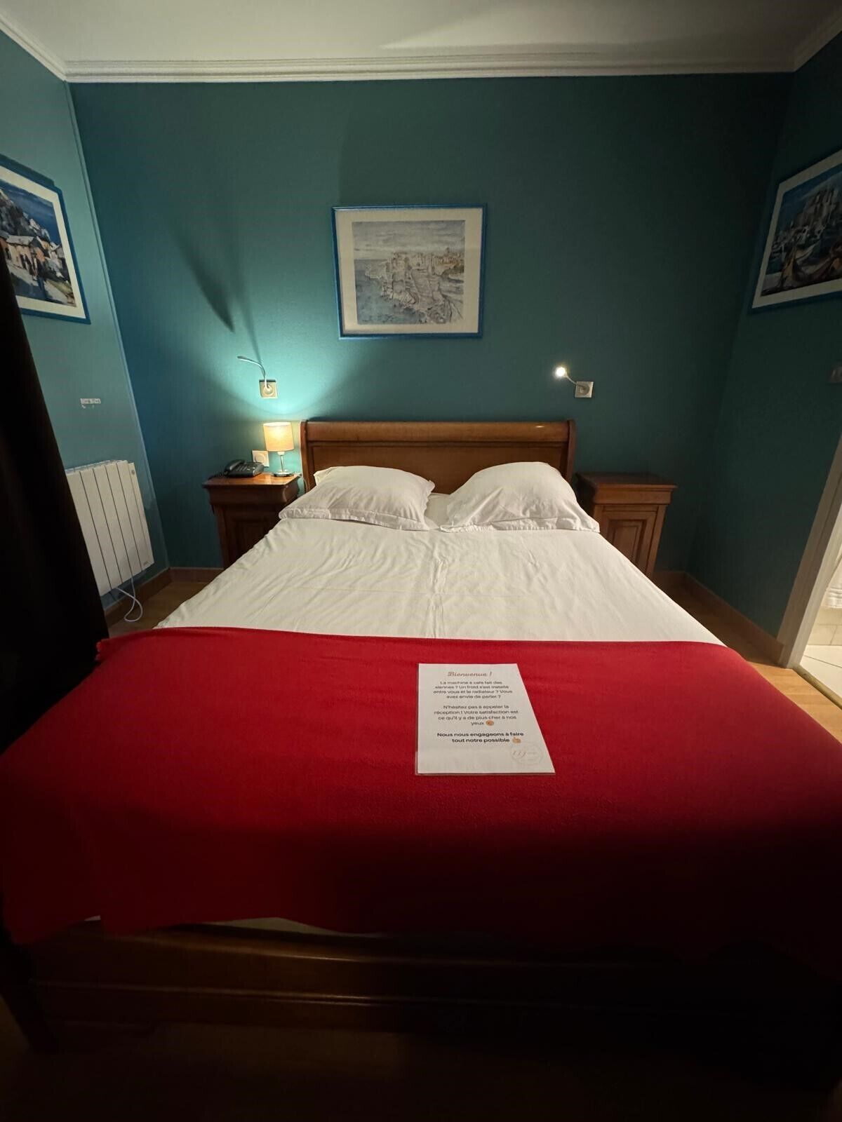 Un lit bien fait, recouvert d’une couverture rouge, avec un tableau encadré au mur et une lampe de chevet.