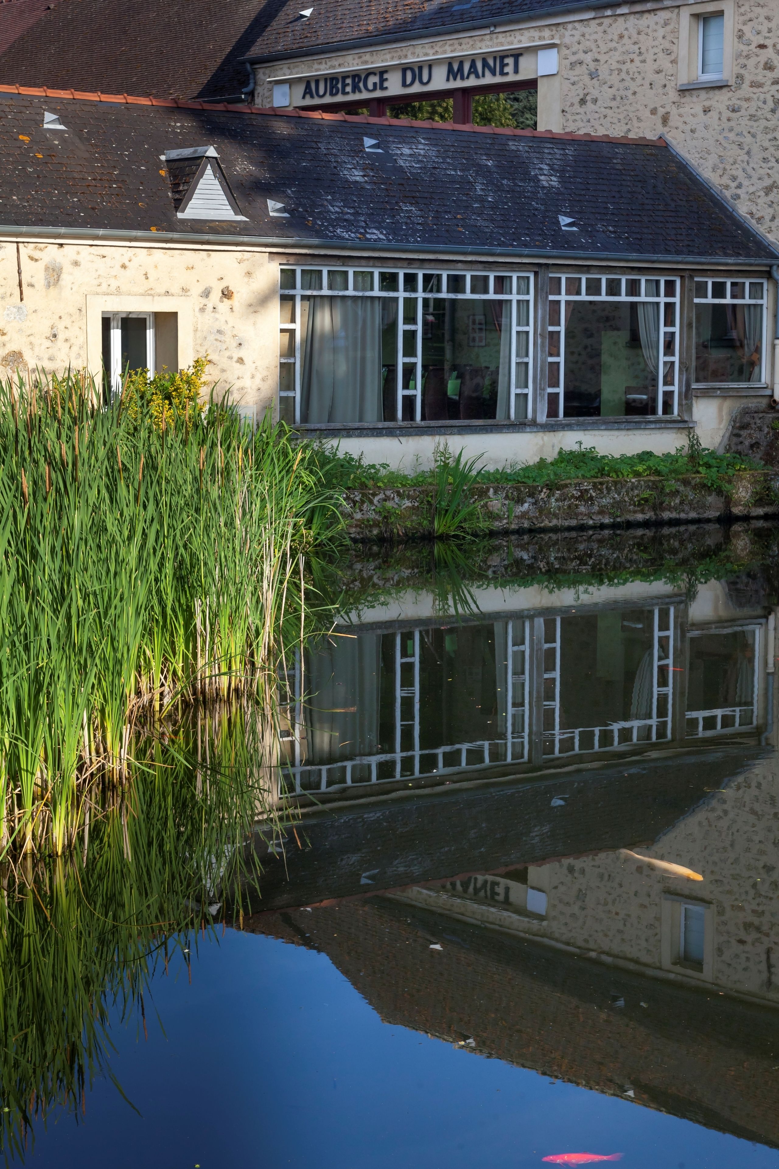 A building with a sign reading "AUBERGE DU MANET" reflecting in a calm water body.