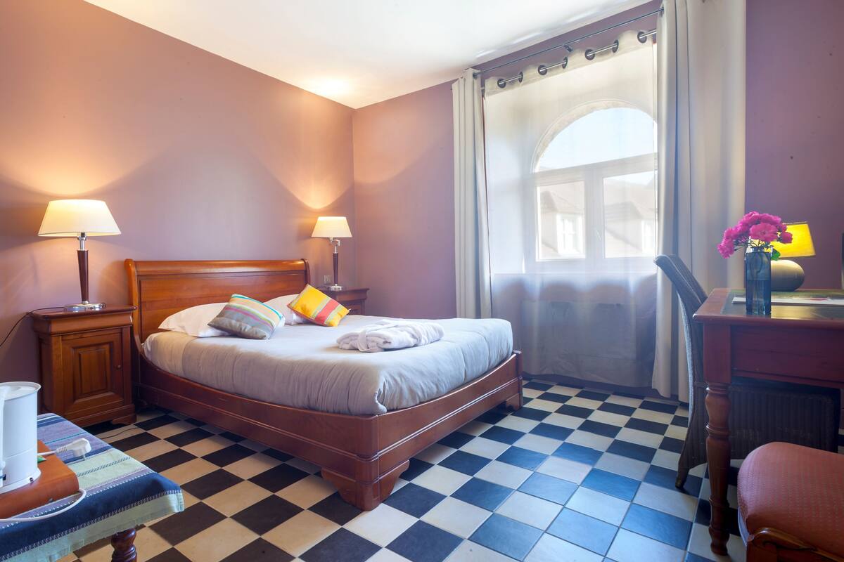 Une chambre d’hôtel avec un sol à carreaux, un lit en bois avec des oreillers, deux lampes de chevet, un bureau avec un vase de fleurs et une fenêtre avec des rideaux.