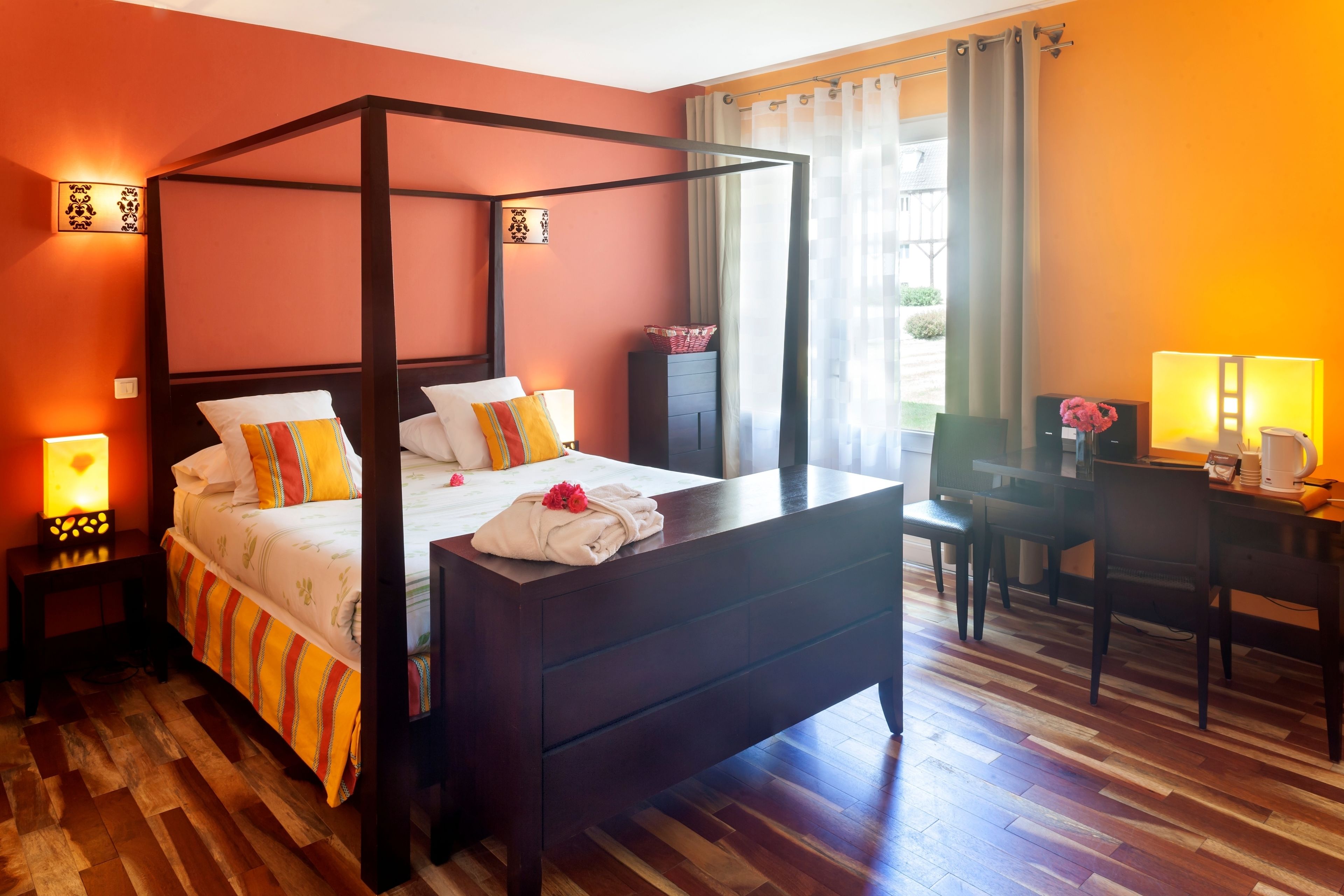 A four-poster bed with a wooden frame, a bedside table with a lamp, a desk with a chair, and a view of the outdoors through a window with curtains.