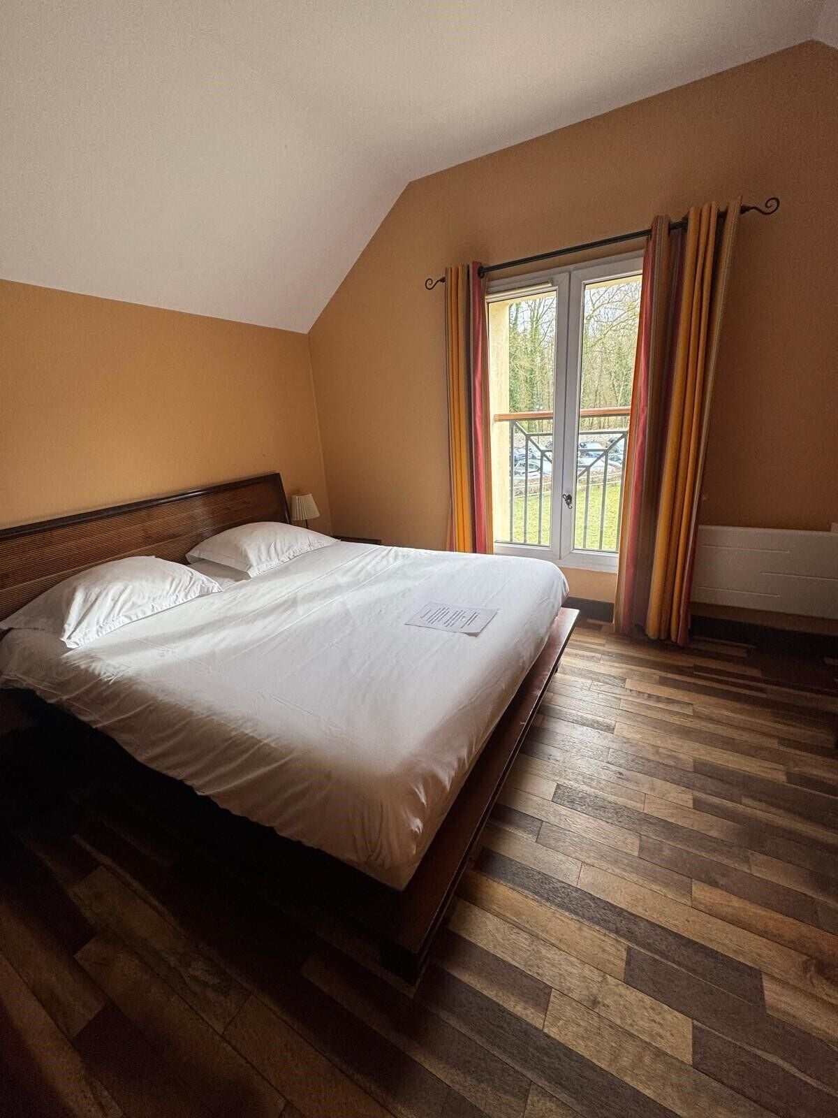 Une chambre avec un plancher en bois, un grand lit, une fenêtre avec des rideaux et une vue sur l’extérieur.