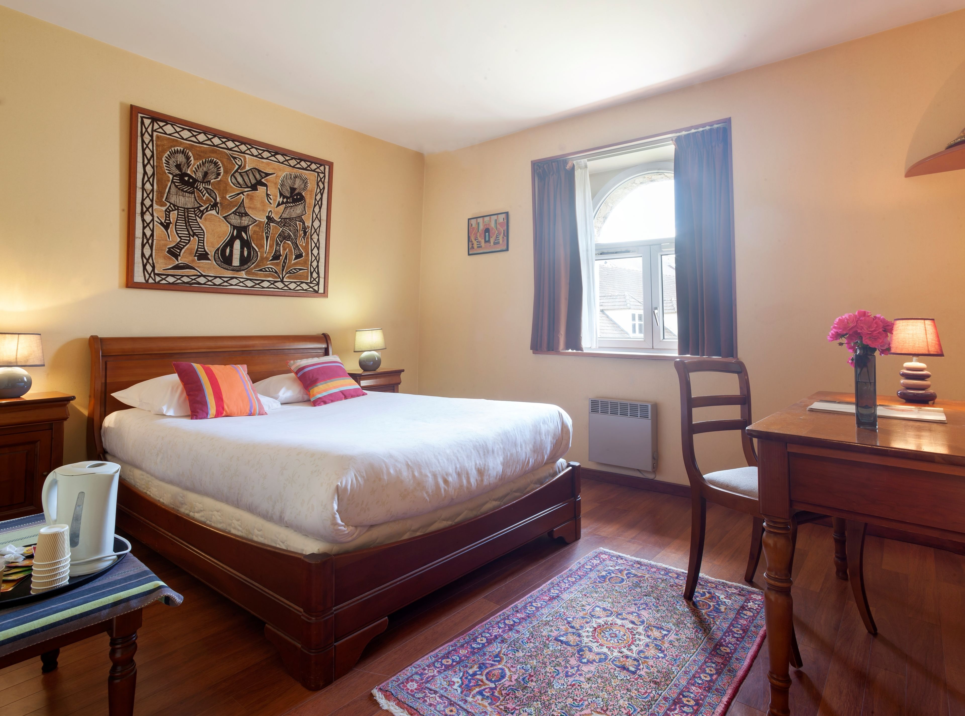 Habitación de hotel ordenada con una cama grande, un escritorio, una silla, una ventana con cortinas, un radiador y un adorno de pared.