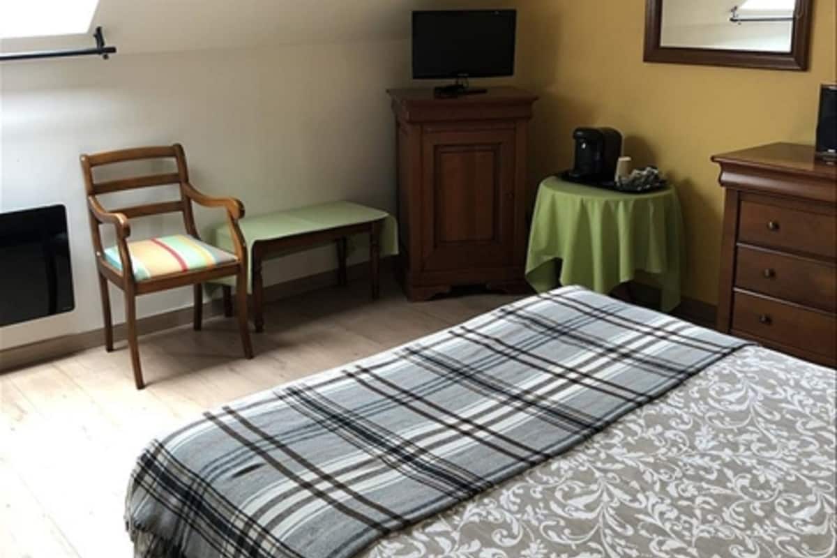 Une chambre à coucher avec un lit, une chaise, un téléviseur posé sur un meuble, une petite table recouverte d’une nappe verte et un miroir accroché au mur.