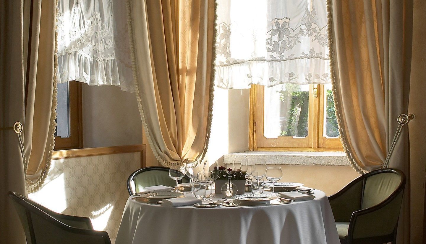 Una mesa de comedor para cuatro personas, con mantel blanco, dos sillas y una ventana con cortinas.