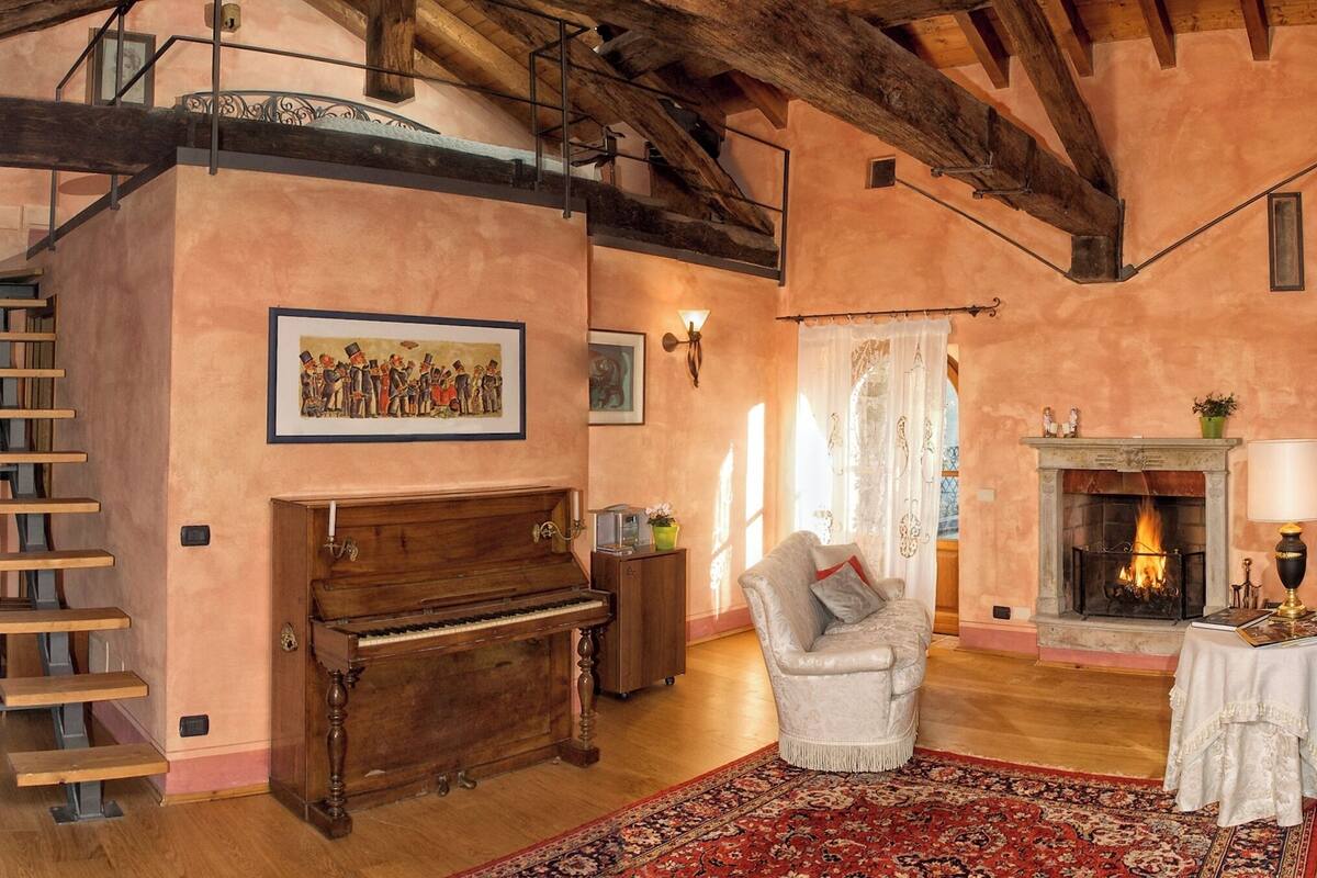A spacious room with a wooden staircase, a grand piano, a comfortable armchair, a fireplace, and a large painting on the wall.