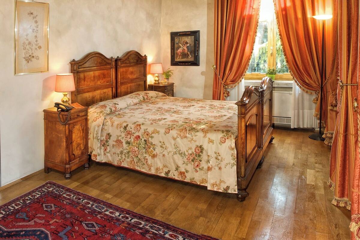 A bedroom with a wooden bed, a nightstand with a lamp, a framed picture on the wall, and a window with curtains.