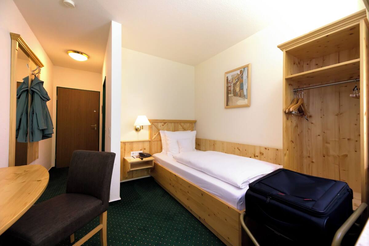Une petite chambre d’hôtel avec un lit simple, une armoire en bois, une chaise, une petite table et une lampe.