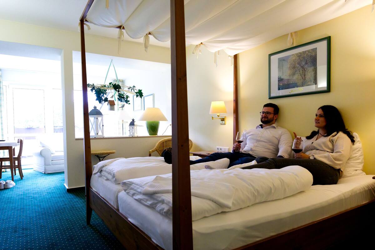 Un lit à baldaquin avec du linge de lit blanc, une petite table avec une lampe et une chaise dans une pièce dont le sol est recouvert d’une moquette bleue.