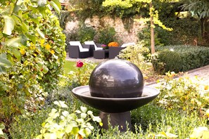 A garden with a black spherical fountain, lush greenery, and outdoor seating with cushions. - Parkhotel Hammerberg (Stolberg)