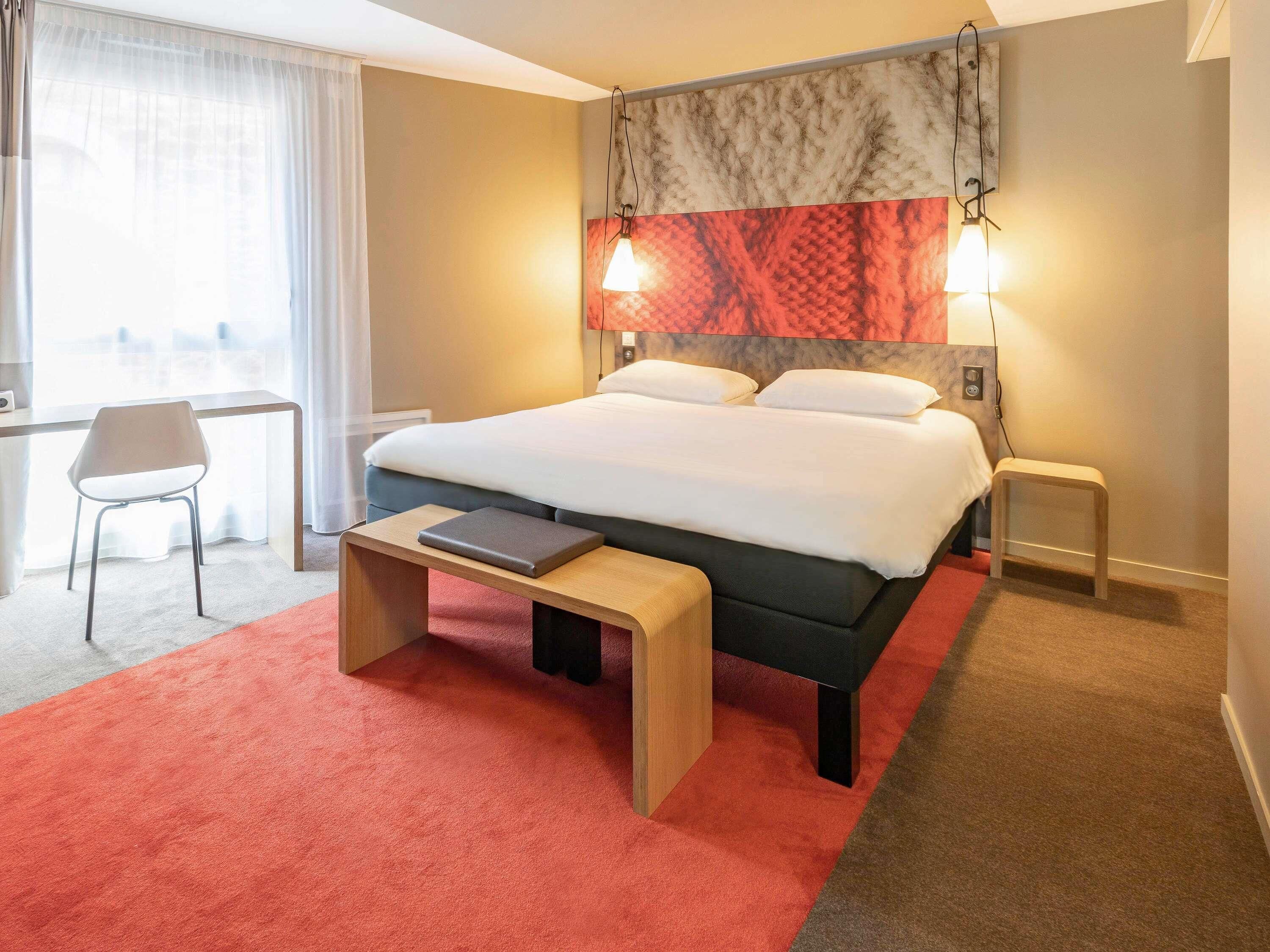 A hotel room with a bed, bedside tables, a desk, a chair, and a view of the city.