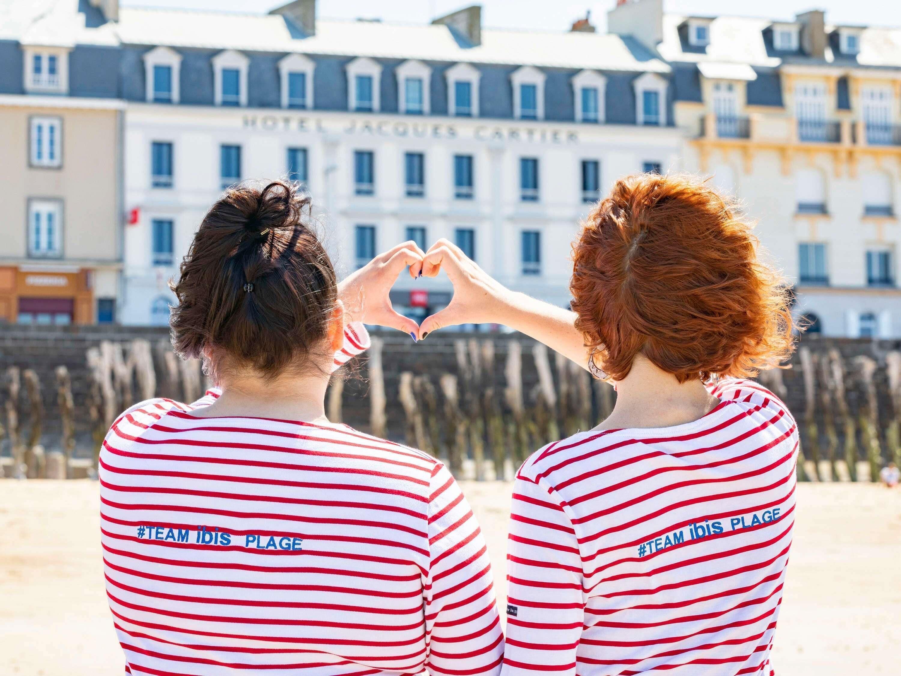 Two people making a heart shape with their hands, wearing matching striped shirts with text, standing in front of a building labeled 'HOTEL JACQUES CARTIER'.