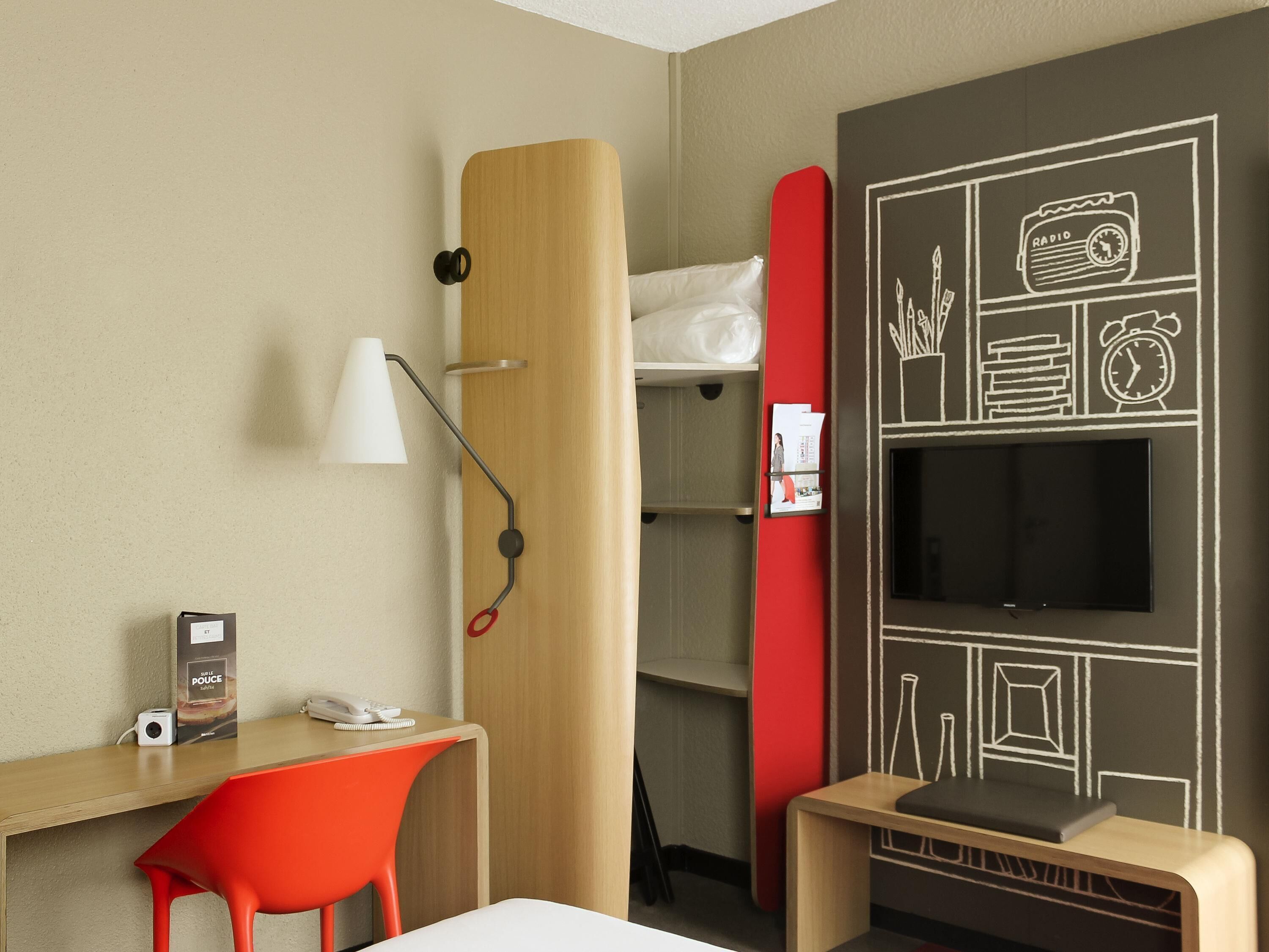 Ein Hotelzimmer mit Schreibtisch, einem roten Sessel, einem Bett mit weißem Kopfkissen, einem Fernseher und einer Wand mit Tafelbild-Dekorationen.