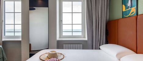 Habitación de hotel con una cama grande, dos ventanas con cortinas, vista al mar, un sombrero y una toalla a rayas sobre la cama.