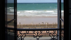 Une vue à travers une fenêtre donnant sur une plage avec des vagues, un ciel dégagé et une balustrade en pierre.