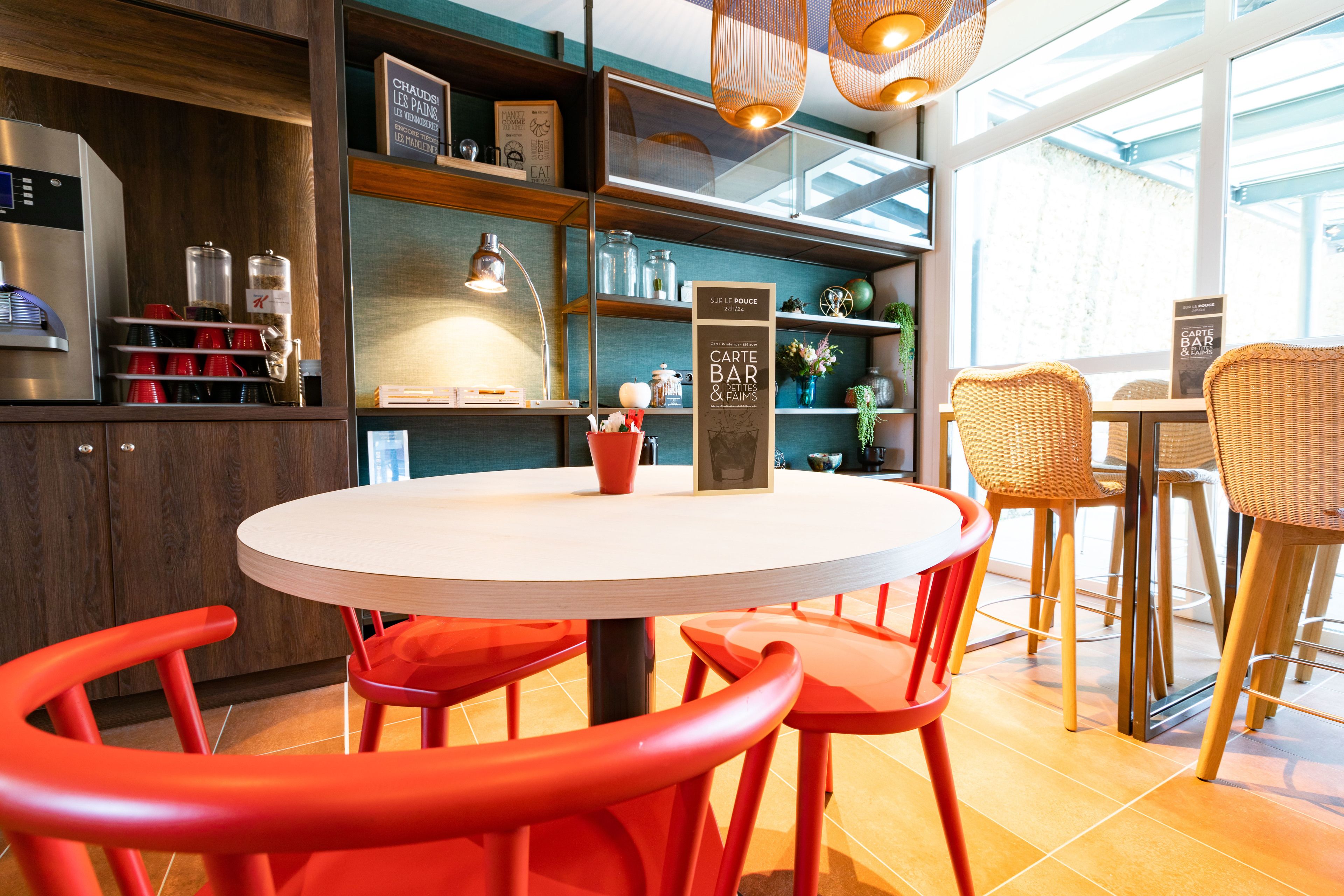 A modern dining area with a round table, red chairs, and wicker chairs. There is a coffee machine and shelves with various items.