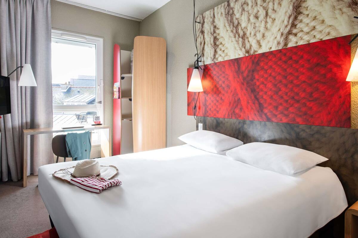 Une chambre d’hôtel avec un lit, un bureau, une chaise, une télévision et une fenêtre avec des rideaux.