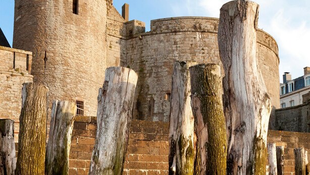Un château de pierre historique, doté d’une tour ronde et d’un grand poteau en bois en premier plan.