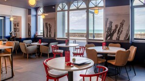 Un café moderne avec des tables rondes et des chaises rouges, de grandes fenêtres donnant sur la mer et des œuvres d’art accrochées aux murs.
