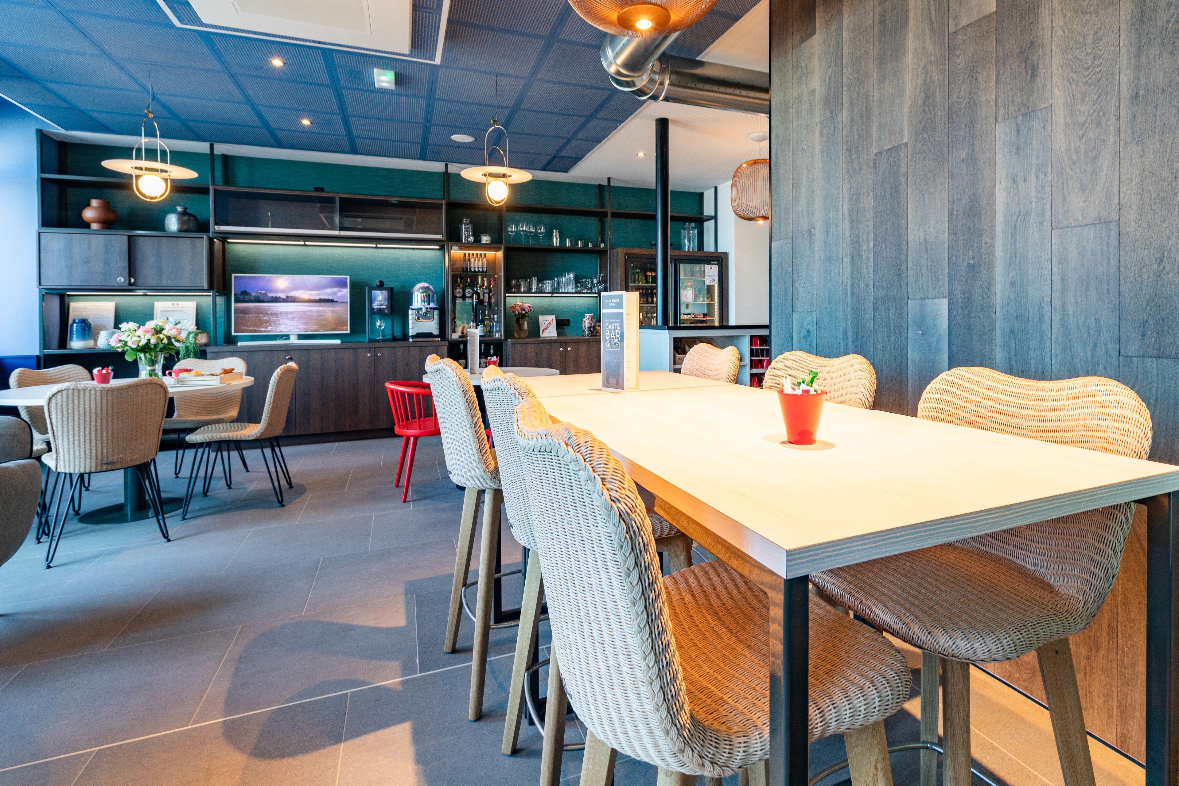 Un comedor moderno con una mesa larga, sillas de mimbre y una silla roja. Hay un televisor montado en la pared y una zona de bar con estantes y botellas.